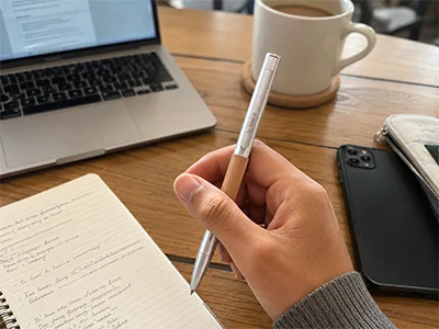 A hand writing in a notebook with the Scriben pen while a laptop sits closed alongside