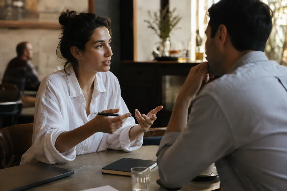 Two people talking across a café table after a meeting, one holding a pen mid-sentence with the laptop closed beside them